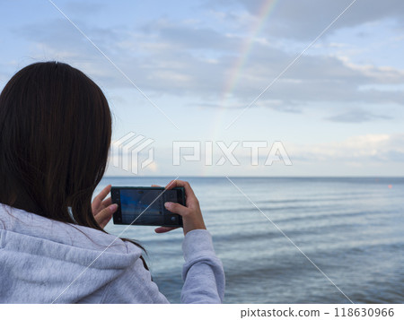 On the shore, young brunette woman aims her smartphone at rainbow taking pictures of its vivid hues. With each snap, her smartphone preserves beauty of moment, capturing the rainbow's essence. On the shore, young brunette woman aims her smartphone at rainbow taking pictures of its vivid hues. With each snap, her smartphone preserves beauty of moment, capturing the rainbow's essence. 118630966
