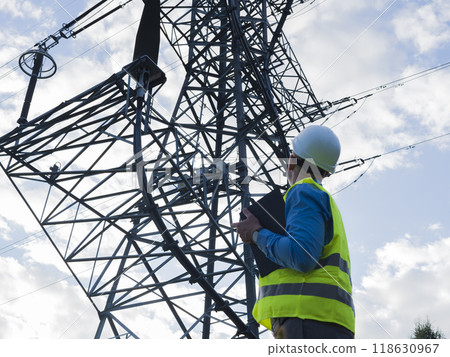 Dedicated energy engineer wearing white helmet and reflective vest holding tablet, observes an electric tower and wires. This energy engineer works to maintain and manage power infrastructure. 118630967