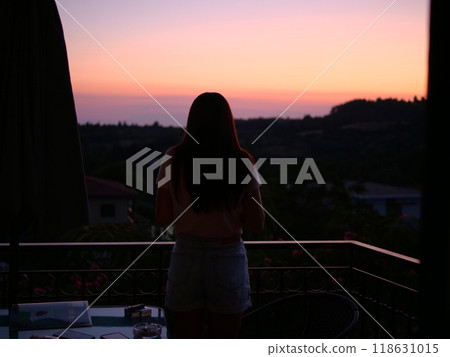 The woman stands with her back and looks at sunset absorbing serene beauty. As the sun sinks below horizon, sunset bathes her in warm hues mesmerizing sunset creates calm and picturesque atmosphere. 118631015