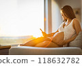Pregnancy concept. Pregnant woman using phone app for online pregnancy prenatal class reading about motherhood or birth touching belly at sunset. Expectant mother relaxing holding belly. 118632782