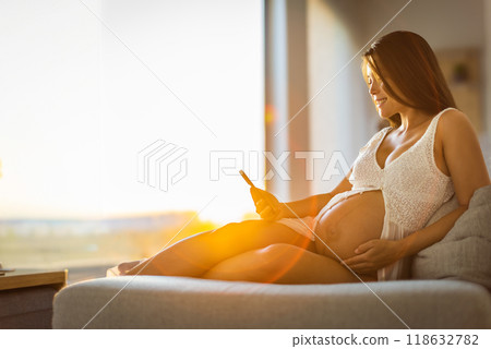 Pregnancy concept. Pregnant woman using phone app for online pregnancy prenatal class reading about motherhood or birth touching belly at sunset. Expectant mother relaxing holding belly. Pregnancy concept. Pregnant woman using phone app for online pregnancy prenatal class reading about motherhood or birth touching belly at sunset. Expectant mother relaxing holding belly. 118632782