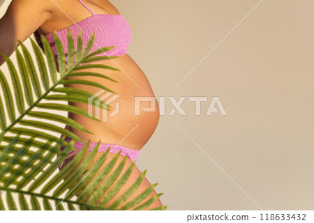 Pregnant woman in pink bikini with belly exposed, tropical leaves in foreground. Pregnancy concept. Pregnant woman in pink bikini with belly exposed, tropical leaves in foreground. Pregnancy concept. 118633432