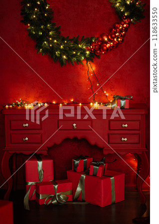 Festive living room interior in red colors with Christmas decorations, boxes with gifts and a wreath on the wall. 118633550