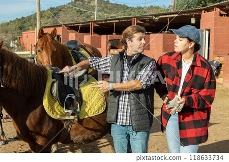 Two skilled man woman stable workers in plaid shirts standing close to saddled horse and discussing upcoming work in countryside club 118633734