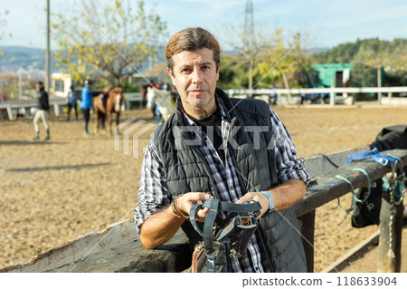 Portrait of skilled adult male stable worker holding horse halter or harness near wooden fence on ranch 118633904