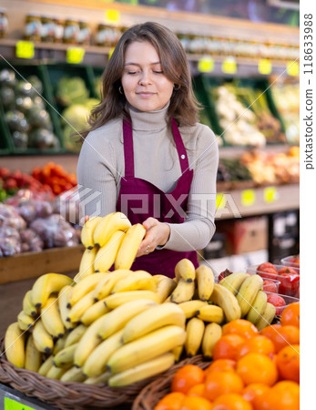 Positive female seller in apron displaying assortment of bananas at supermarket 118633988
