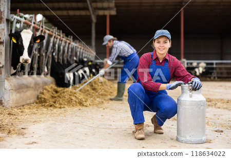 Cheerful hardworking woman farmer carrying milk can while walking through cow house in autumn 118634022