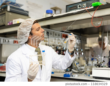 Serious Latin male biochemist holding tripod and examining solution in test tube for chemical reaction in research center 118634329