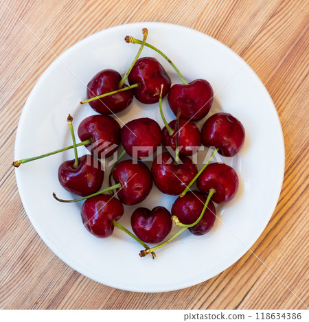 There is plate with ripe maroon cherries on simple wooden table. There is plate with ripe maroon cherries on simple wooden table. 118634386