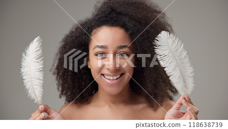 Health, beauty, tenderness and softness concept - beautiful African American woman with curly afro hairstyle and clean, healthy skin, smiles and touches her face with a white fluffy feather 118638739