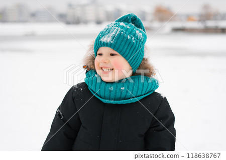 Happy boy wearing a blue hat and scarf is smiling in the snow. Child is wearing a black coat and he is enjoying the winter weather Happy boy wearing a blue hat and scarf is smiling in the snow. Child is wearing a black coat and he is enjoying the winter weather 118638767