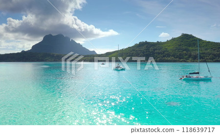 Sailboats anchored in turquoise lagoon, green mountains tropical island in background. Crystal clear water reflects blue sky. Yacht catamaran boat tour to French Polynesia. Exotic summer luxury travel 118639717
