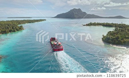 Red cargo ship sailing turquoise water lagoon, passing near tropical island with lush vegetation palm trees beach. Delivery bulk carrier to exotic remote places, Maupiti, French Polynesia. Aerial view 118639725
