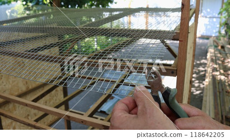 worker's hands with nippers cutting wire mesh during assembly of quail cage, construction of equipment for home keeping of small laying hens, work with hand tool creating cage 118642205