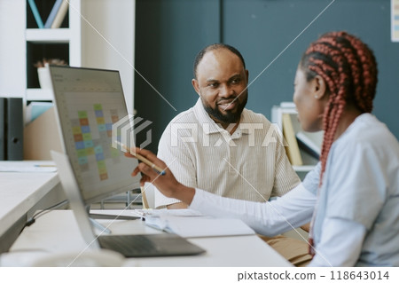 Two professionals discussing work tasks at an office desk while viewing digital charts on a monitor Clients or colleagues collaborating and sharing ideas in workplace 118643014