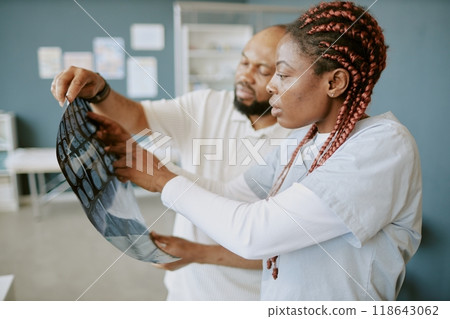 Two African American medical professionals examining X-rays in clinical setting, discussing patient health while wearing lab coats, with healthcare posters visible in background 118643062