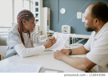 Healthcare professional consulting with patient, discussing medical records in clinic room with shelves and books in background Healthcare professional consulting with patient, discussing medical records in clinic room with shelves and books in background 118643090
