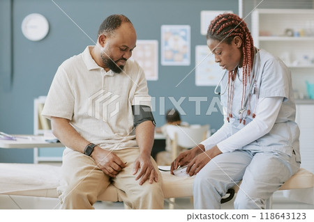 Medical professional in uniform with stethoscope examining patient seated in clinic setting providing care and attention against clinical backdrop Medical professional in uniform with stethoscope examining patient seated in clinic setting providing care and attention against clinical backdrop 118643123