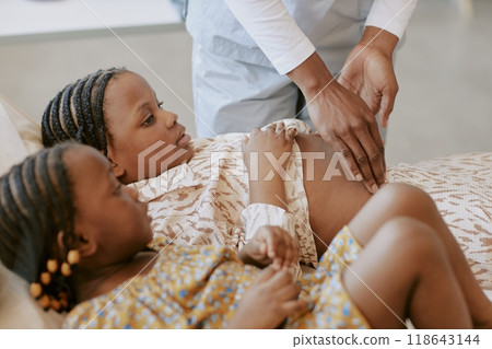 Medical professional examining an African childs abdomen on a bed while another child watches. Gentle interaction during healthcare check-up creating a caring environment Medical professional examining an African childs abdomen on a bed while another child watches. Gentle interaction during healthcare check-up creating a caring environment 118643144