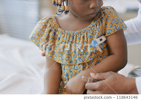 African American girl with arm thermometer sitting on bed while caregiver showing concern and providing comfort in softly lit room during healthcare check-up African American girl with arm thermometer sitting on bed while caregiver showing concern and providing comfort in softly lit room during healthcare check-up 118643146
