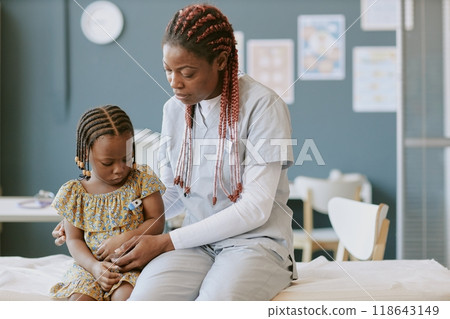 Medical professional in scrubs examining young child's arm in clinical setting. Child wearing dress while doctor attentively checks her health in bright, organized room Medical professional in scrubs examining young child's arm in clinical setting. Child wearing dress while doctor attentively checks her health in bright, organized room 118643149