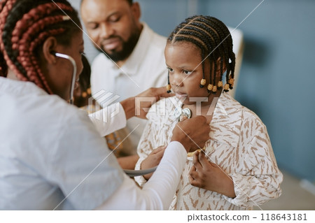 Child receiving medical examination with stethoscope by professional medical practitioner with other adults in background looking with concern 118643181