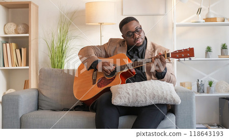Guitar practice. Musician leisure. Talented peaceful man playing song chords on string instrument enjoying creative activity on couch in living room at home. Guitar practice. Musician leisure. Talented peaceful man playing song chords on string instrument enjoying creative activity on couch in living room at home. 118643193