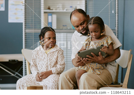 Biracial father spending quality time with daughters teaching them using tablet in living room creating a bonding moment while sitting together on chairs 118643211