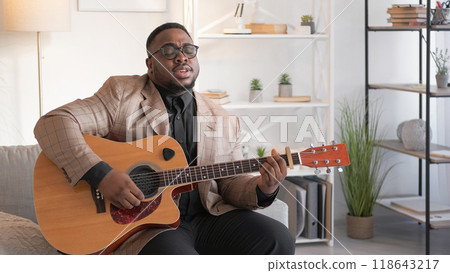 Song performer. Male vocalist. Musical hobby. Talented inspired man artist singing melody playing acoustic guitar on couch at home in living room. Song performer. Male vocalist. Musical hobby. Talented inspired man artist singing melody playing acoustic guitar on couch at home in living room. 118643217