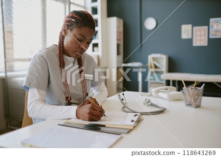 Nurse seated at desk in medical office reviewing patient records and taking notes. Medical equipment including stethoscope and telephone visible on desk Nurse seated at desk in medical office reviewing patient records and taking notes. Medical equipment including stethoscope and telephone visible on desk 118643269