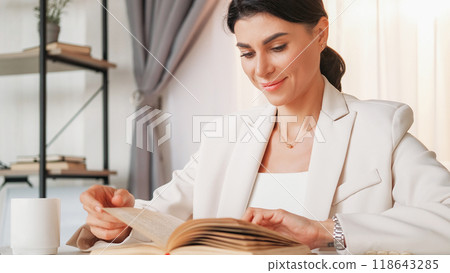 Enjoying book. Happy woman. Work break. Elegant smiling lady sitting office desk reading interesting literature in light room interior. 118643285