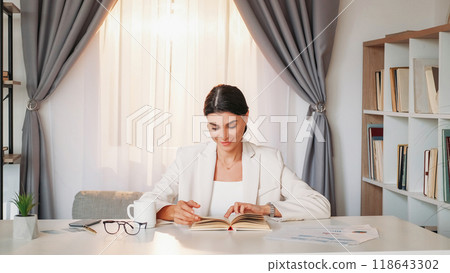 Rest work. Happy woman. Office lifestyle. Elegant smiling lady sitting desk reading interesting book in light room interior. Rest work. Happy woman. Office lifestyle. Elegant smiling lady sitting desk reading interesting book in light room interior. 118643302