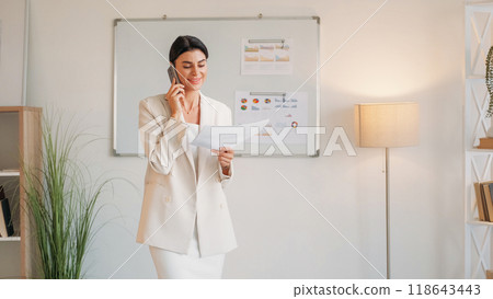 Business process. Elegant woman. Mobile chatting. Smiling female chief talking smartphone holding paper documents light room interior. Business process. Elegant woman. Mobile chatting. Smiling female chief talking smartphone holding paper documents light room interior. 118643443