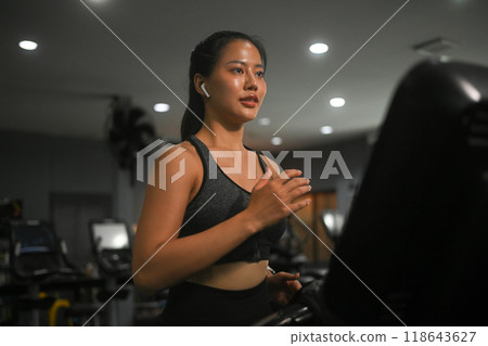 Young sporty woman listening to music and running on a treadmill at gym. Sports and healthy lifestyle concept Young sporty woman listening to music and running on a treadmill at gym. Sports and healthy lifestyle concept 118643627
