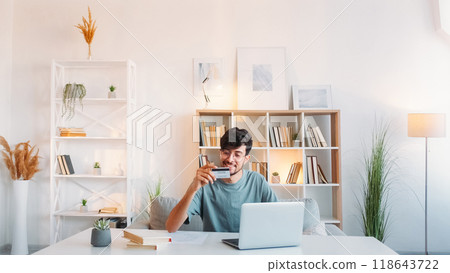 Online banking. Internet payment. Electronic transaction. Satisfied smiling man using credit card laptop at light modern home workplace interior with free space. Online banking. Internet payment. Electronic transaction. Satisfied smiling man using credit card laptop at light modern home workplace interior with free space. 118643722