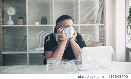 Pandemic isolation. Office quarantine. Bored tired unhappy overweight woman in medical mask during business shutdown in protected plastic workspace. 118643790