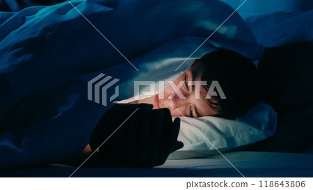Guy using smartphone under bedsheets at night in dim light indicating addiction insomnia in bed at night. 118643806