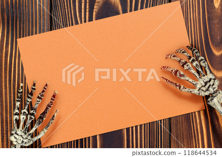 A human skeleton hand holding an orange piece of paper on a wooden background. The blank orange piece of paper for copy space. The image has a creepy and unsettling mood, a horror A human skeleton hand holding an orange piece of paper on a wooden background. The blank orange piece of paper for copy space. The image has a creepy and unsettling mood, a horror 118644534