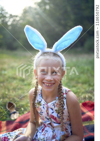 little blonde girl with braids wearing an easter headpiece with bunny ears on a picnic in the park little blonde girl with braids wearing an easter headpiece with bunny ears on a picnic in the park 118650792