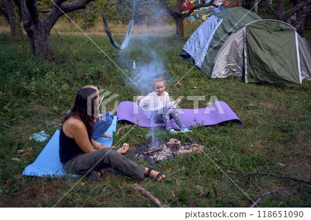 mother and her daughters roasting and eating marshmallows on the campfire, camping vacation with children, mosquito bites on children's faces mother and her daughters roasting and eating marshmallows on the campfire, camping vacation with children, mosquito bites on children's faces 118651090