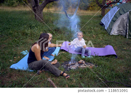 mother and her daughters roasting and eating marshmallows on the campfire, camping vacation with children, mosquito bites on children's faces mother and her daughters roasting and eating marshmallows on the campfire, camping vacation with children, mosquito bites on children's faces 118651091