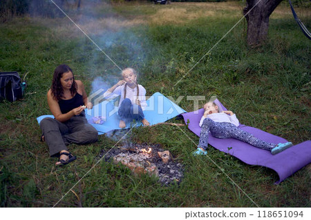 mother and her daughters  roasting and eating marshmallows on the campfire, camping vacation with children, mosquito bites on children's faces 118651094