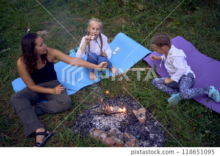 mother and her daughters roasting and eating marshmallows on the campfire, camping vacation with children, mosquito bites on children's faces mother and her daughters roasting and eating marshmallows on the campfire, camping vacation with children, mosquito bites on children's faces 118651095
