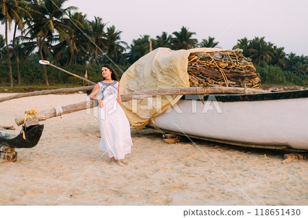 Goa, India. Young Caucasian Woman In White Dress Standing Near Fishing Boat And Posing For Camera Goa, India. Young Caucasian Woman In White Dress Standing Near Fishing Boat And Posing For Camera 118651430