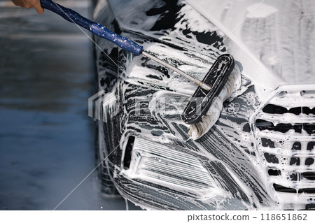 Man washes a car using a special brush at a car wash. Close up of man wash right headlight. The car is in foam Man washes a car using a special brush at a car wash. Close up of man wash right headlight. The car is in foam 118651862