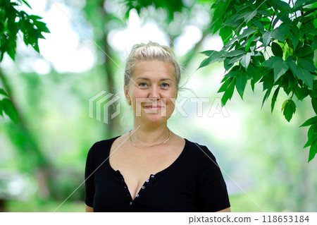 Real people, attractive simple woman in her 30's with first wrinkles on her face looks serious, standing in a park under green leaves in summer. Real people, attractive simple woman in her 30's with first wrinkles on her face looks serious, standing in a park under green leaves in summer. 118653184