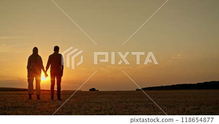 A young farming couple stands in a field, holding hands at sunset, while a combine harvests in the distance. A young farming couple stands in a field, holding hands at sunset, while a combine harvests in the distance. 118654877