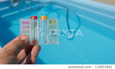 A hand holds a pool water testing kit over a clear blue swimming pool with cleaning tools in the background. 118654882