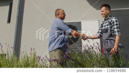 A technician and homeowner finalize their consultation with a handshake beside an external heat pump unit. A technician and homeowner finalize their consultation with a handshake beside an external heat pump unit. 118654886