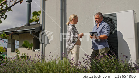 A homeowner reviews heat pump settings on a tablet with a technician outdoors. A homeowner reviews heat pump settings on a tablet with a technician outdoors. 118654890